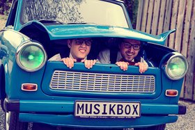 zwei Männer schauen aus dem Kofferraum eines blauen, alten Autos der Marke Trabant.