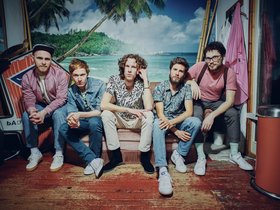 Die fünf Jungs der Band anders sitzen auf einer Couch vor einer Wand mit Hawaii-Tapete. 