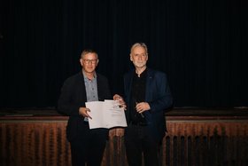 Minski-Programmgestalter Michael Mertes (links) und Kulturstaatssekretär Jürgen Hardeck vor dunklem Hintergund. Mertes präsentiert die Preisurkunde.