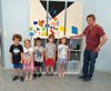 Fünf Kindergartenkinder und ein Mann stehen vor einem Bücherregal, auf dem drei Leinwände, die mit geometrischen Formen bemalt sind,stehen.